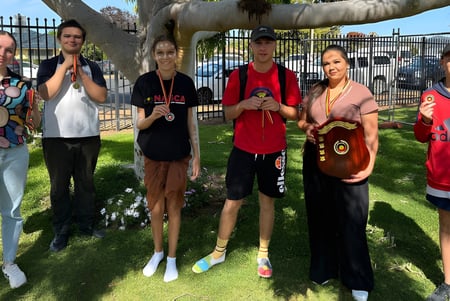 Un grupo de estudiantes y adultos está de pie en un prado en el terreno de la Wellington High School frente a un gran árbol.