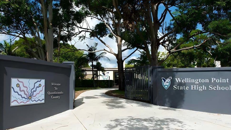 La entrada a la Wellington Point State High School con el cartel del edificio escolar y rodeada de árboles y plantas.