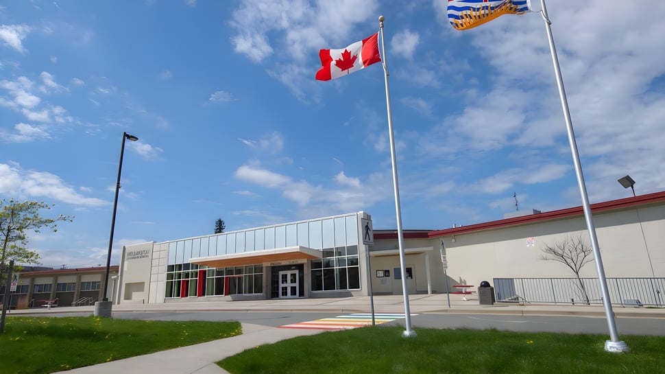 La bandera canadiense ondea sobre el moderno edificio de vidrio de la Wellington Secondary School bajo un cielo azul.