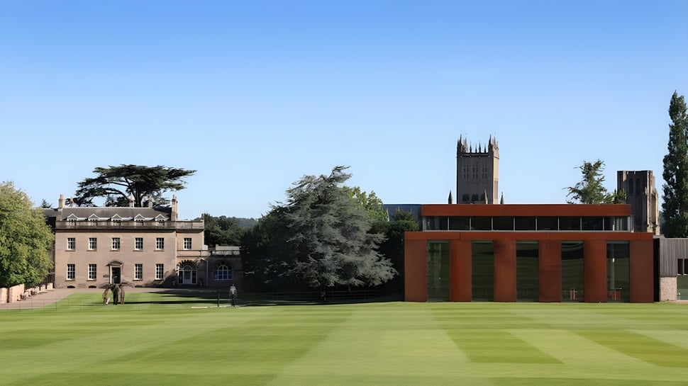El campus verde de la Wells Cathedral School con edificios históricos y una torre moderna al fondo bajo un cielo azul.