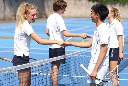Estudiantes de la Wells Cathedral School se dan la mano en una colorida cancha de tenis al aire libre.