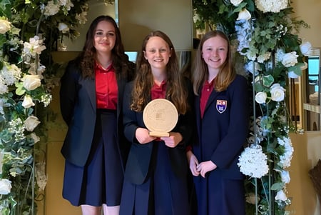 Tres graduadas del Wesley College están juntas frente a un arco decorado con flores y sosteniendo su diploma.