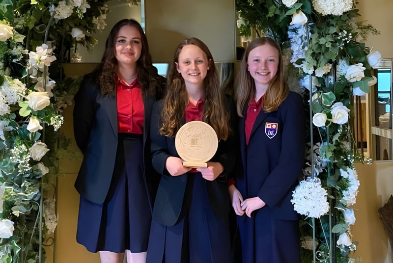 Tres graduadas del Wesley College están juntas frente a un arco decorado con flores y sosteniendo su diploma.