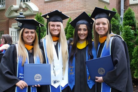 Cuatro graduadas en toga y birrete sostienen sus diplomas frente a un edificio de ladrillo en el campus del Wesley College.