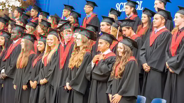 Un gran grupo de graduados de la West Carleton Secondary School está de pie en túnicas negras y estolas rojas frente a un fondo azul con el logo de la escuela.