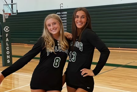 Dos estudiantes de la West Catholic High School están en trajes deportivos en un gimnasio frente a una pared verde y sonríen.