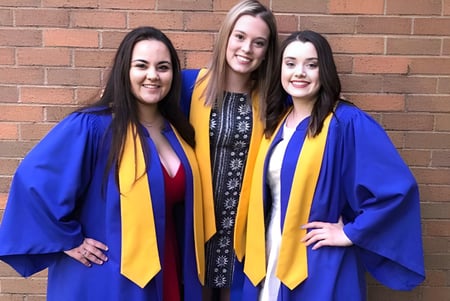 Tres estudiantes en togas de graduación azules y amarillas posan frente a una pared de ladrillos en el campus de la Westisle Composite High School.