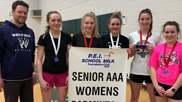 Un grupo de estudiantes de la Westisle Composite High School sostiene una pancarta para el evento Senior AAA Womens de la P.E.I. School Milk Foundation en un gimnasio.