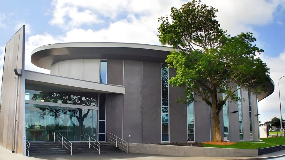 El moderno edificio con fachada de vidrio y un gran árbol en primer plano en el campus de la Westlake Girls High School bajo un cielo nublado.