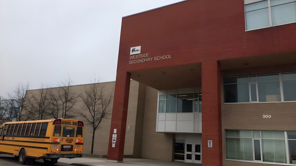 Se puede ver el edificio de ladrillo de la Westside Secondary School con un autobús escolar amarillo delante.