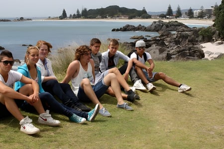 Un grupo de estudiantes de la Whakatane High School está sentado en el césped cerca de la costa rocosa con agua y edificios al fondo.