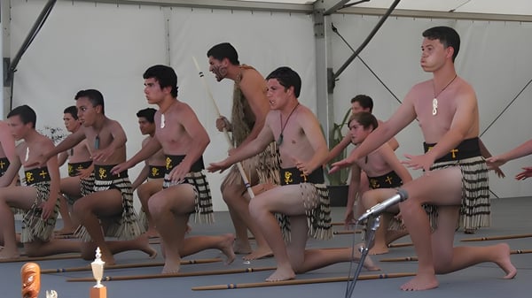 Un grupo de hombres en ropa tradicional realiza un baile sincronizado en el escenario del campus de la Whangamata Area School.