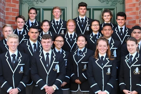 Un grupo de estudiantes de la Whanganui Collegiate School está en uniformes negros uniformes frente a un edificio de ladrillo con elementos decorativos.