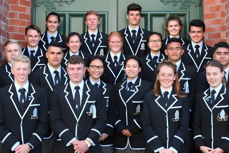 Un grupo de estudiantes de la Whanganui Collegiate School está en uniformes negros uniformes frente a un edificio de ladrillo con elementos decorativos.