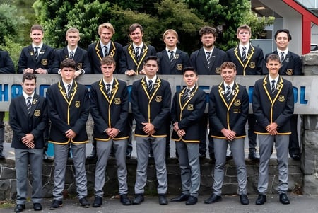 Un grupo de estudiantes de Whangarei Boys’ High School está de pie en uniforme escolar formal frente a un edificio.