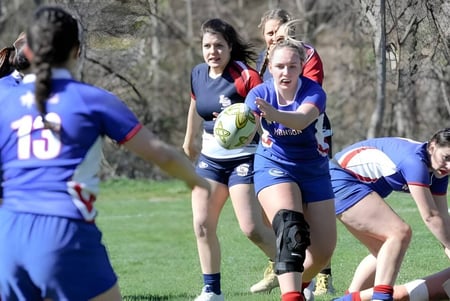 Jugadoras de rugby de la Wilbraham & Monson Academy en un partido en un campo de hierba con árboles al fondo.