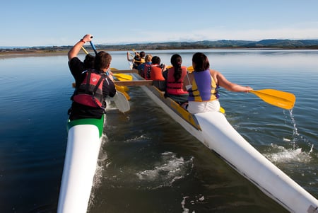 Estudiantes del William Colenso College reman juntos en una canoa de colores en un lago tranquilo con montañas al fondo.