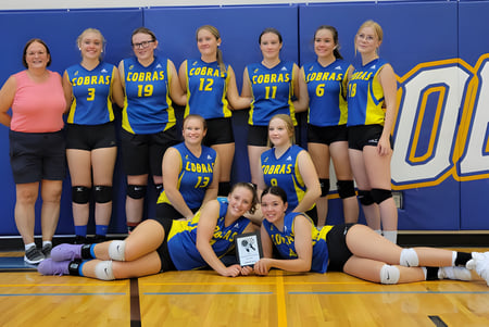 Un equipo de jugadoras de voleibol en la cancha de baloncesto de la Willow Creek Composite High School posa junto a un gran logo de OB.