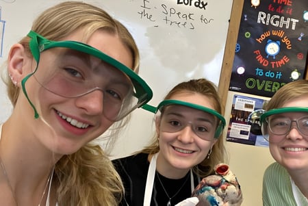 Tres estudiantes de la Winchendon School con gafas de protección están en el aula frente a una pizarra.