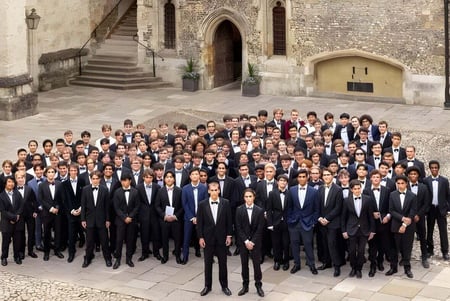 Un gran grupo de estudiantes vestidos de manera formal está frente al edificio histórico de piedra con arcadas decoradas en el campus de Winchester College.