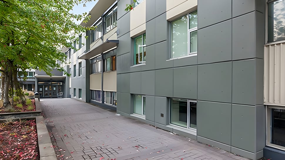 El edificio de varios pisos de la Windsor Secondary School con fachada gris se encuentra en un camino pavimentado y está rodeado de árboles.