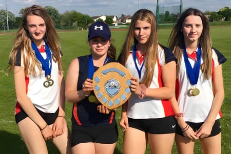 Cuatro estudiantes de la Wisbech Grammar School sostienen medallas y un trofeo en el campo de deportes.