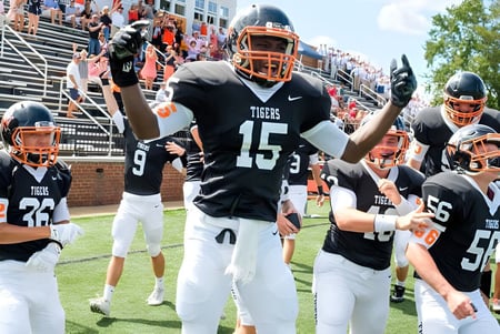 Jugadores de la Woodberry Forest School en camisetas negras y naranjas celebran en el campo frente a los espectadores en las gradas.