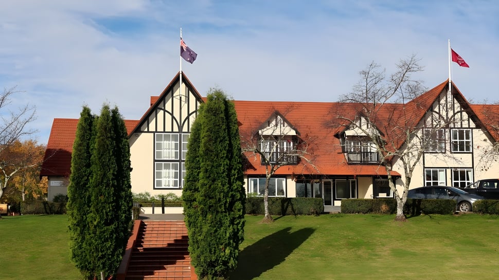 El edificio Tudor de Woodford House con techos de tejas rojas y fachadas de entramado de madera se encuentra en un césped cuidado rodeado de cipreses.