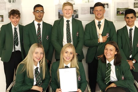 Un grupo de estudiantes de la Woodhouse Grove School en uniforme escolar verde posan en un edificio escolar frente a imágenes en la pared.