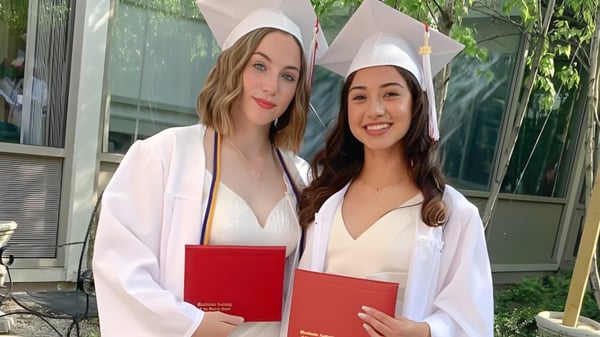 Dos estudiantes de la Woodlands Academy of the Sacred Heart en ropa de graduación sostienen orgullosamente sus diplomas frente a un edificio con área verde.