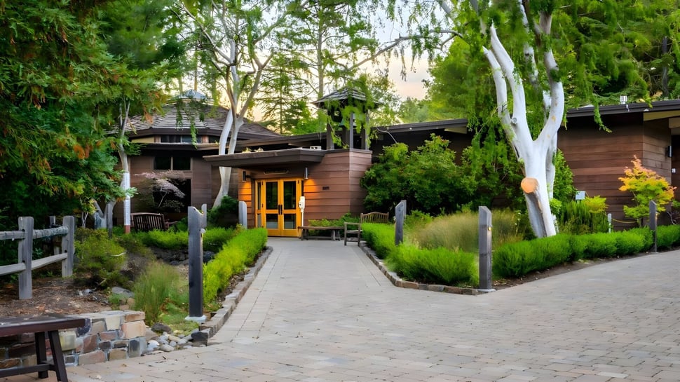 Un edificio de madera rústico con una entrada acogedora se encuentra en el terreno de la Woodside Priory School rodeado de árboles y un camino de piedra.