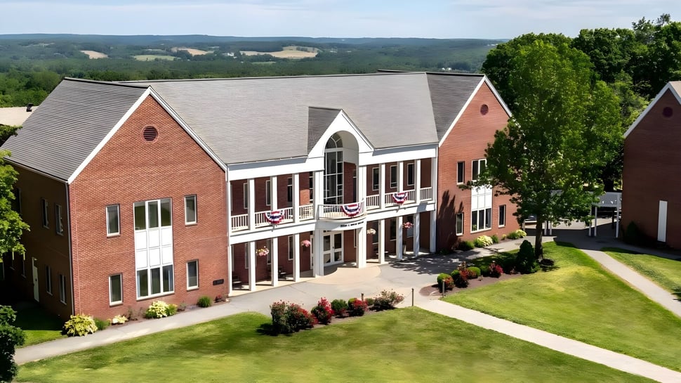 El edificio de ladrillo de varios pisos de la Woodstock Academy se encuentra frente a colinas verdes y rodeado de vegetación exuberante.