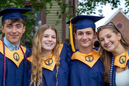 Cuatro estudiantes en togas de graduación posan juntos frente a un edificio en el campus de la Woodstock Academy.