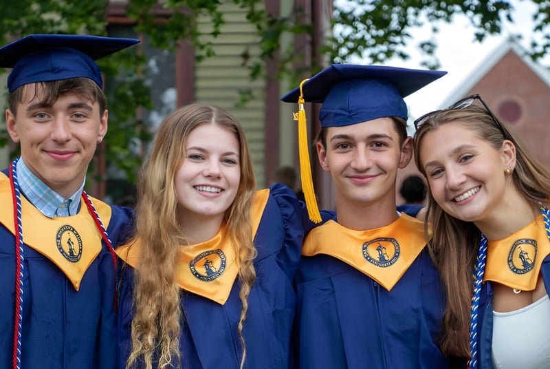 Cuatro estudiantes en togas de graduación posan juntos frente a un edificio en el campus de la Woodstock Academy.
