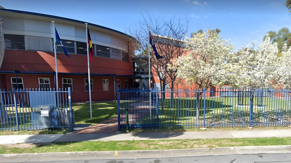 El moderno edificio principal de varios pisos de la Woodville High School con techo arqueado se encuentra frente a un área verde cercada y árboles en flor.
