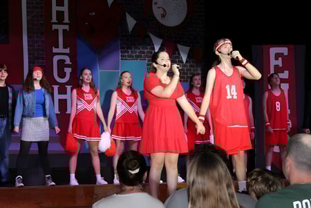 Un grupo de estudiantes de la Woolgoolga High School se presenta en trajes de animadoras rojos en un escenario con varios carteles Públicoitarios al fondo.