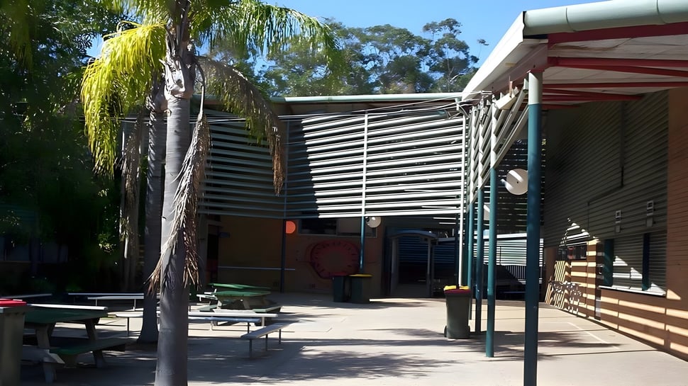 El patio de recreo de la Woolgoolga High School muestra una zona de asientos sombreada con bancos de madera y mucho verde.