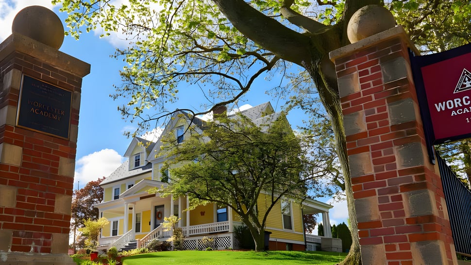 Un arco de ladrillo enmarca una calle arbolada con una casa amarilla y un edificio de ladrillo rojo en el terreno de la Worcester Academy.