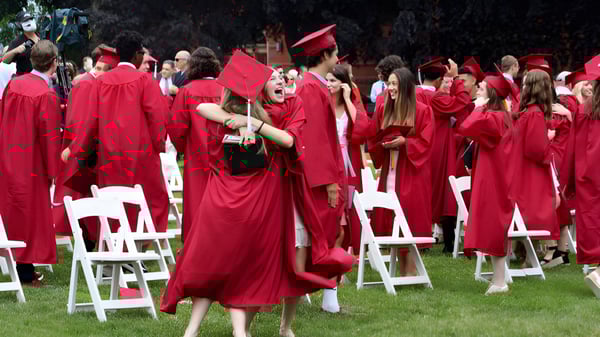 Un gran grupo de graduados y graduadas de la Worcester Academy está de pie en túnicas y sombreros rojos en un campo de hierba frente a un escenario.