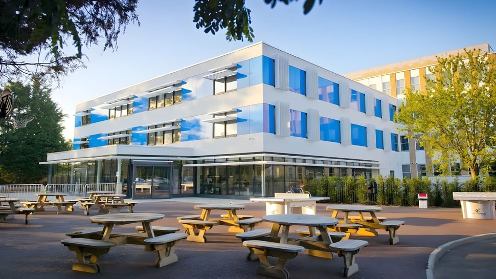 El moderno edificio de varios pisos con fachada de vidrio de la Worthgate School Canterbury está rodeado de un área exterior con mesas de madera.