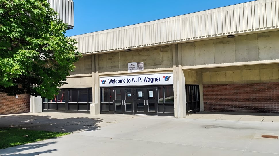 El edificio de la W.P. Wagner High School con un cartel de bienvenida sobre la entrada está rodeado de árboles y áreas verdes.