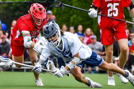 Dos jugadoras en rojo y blanco juegan lacrosse en el campo de Wyoming Seminary College Preparatory con espectadores al fondo.