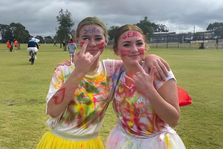 Dos alumnas del Yanchep Secondary College llevan camisetas de Tie-Dye coloridas y hacen muecas al aire libre con gestos de las manos.